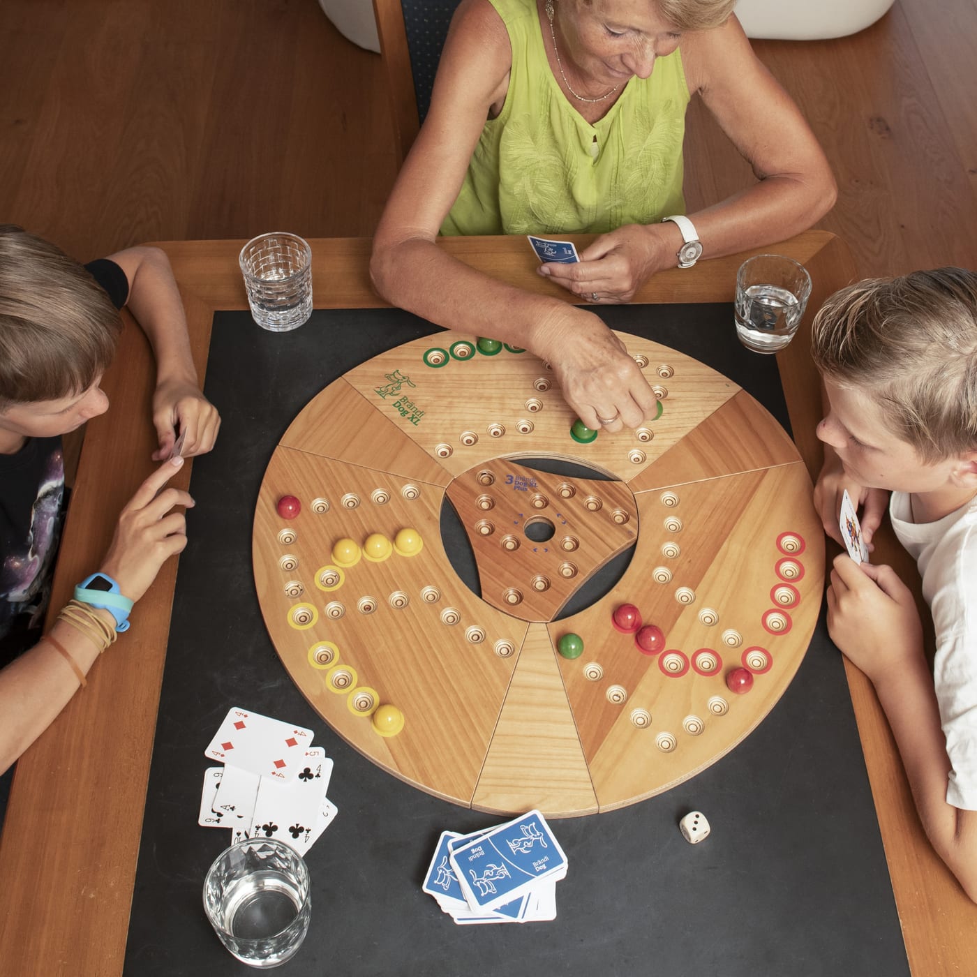 Drei Personen spielen Brändi Dog XL am Tisch, rundes Spielbrett mit grossen Murmeln, daneben Karten und Würfel